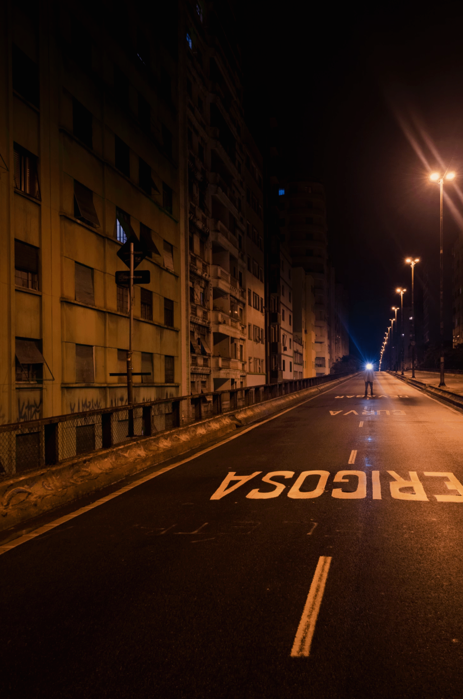 A human figure with a blue light emanating from its head moves towards the camera through an elevated highway illuminated by dim orange light poles